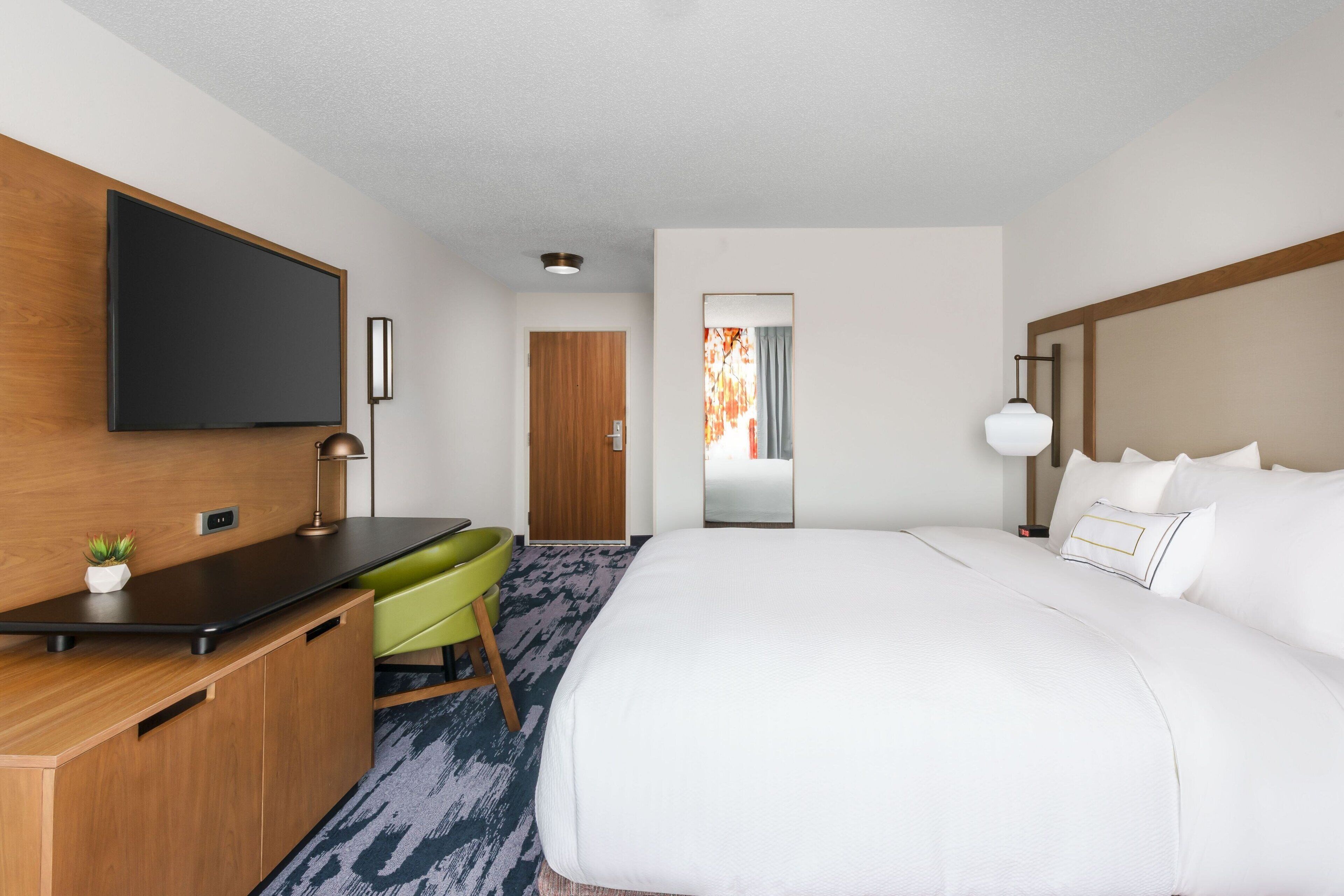 A hotel room with a large bed, a desk, a chair, a flat-screen TV mounted on the wall, a wooden door, and a window with curtains.