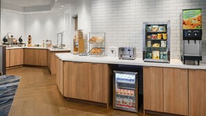Un área de cocina moderna con salpicadero blanco de azulejos, armarios de madera, un refrigerador de acero inoxidable, una cafetera y un expositor para pan.