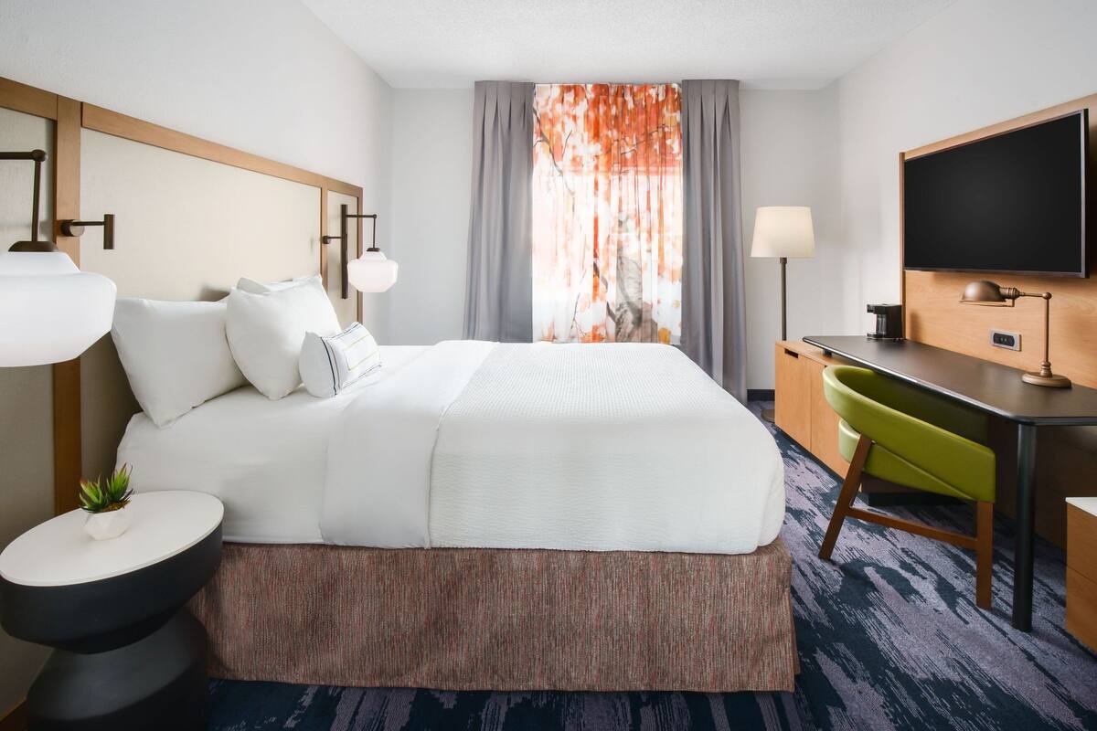 A hotel room with a large bed, a desk, a chair, a TV, and a window with curtains.