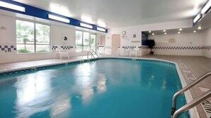 Una piscina interior con agua azul cristalina, rodeada de sillas reclinables blancas y un suelo de baldosas.