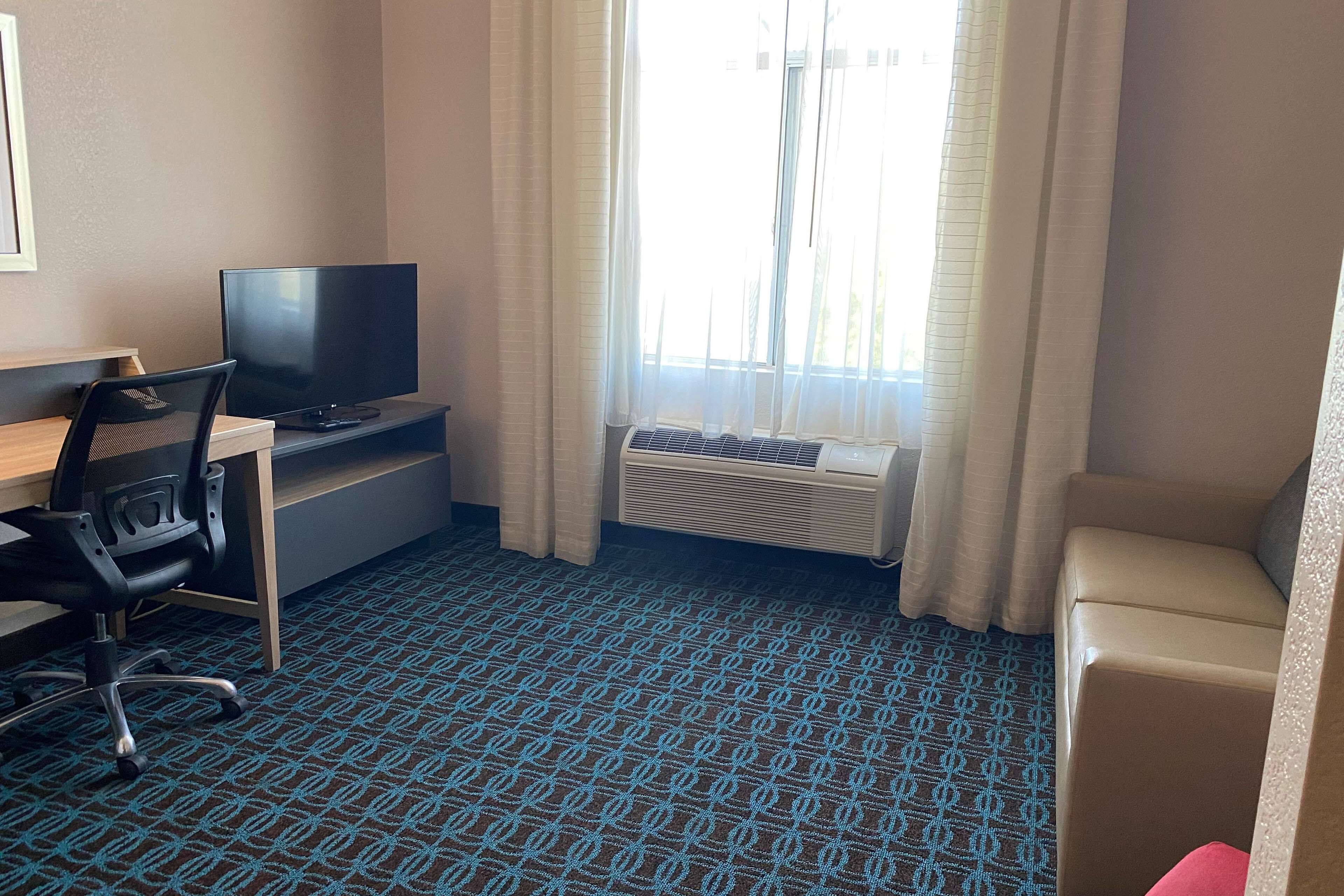 A hotel room with a desk, chair, television, air conditioning unit, and a window with curtains.