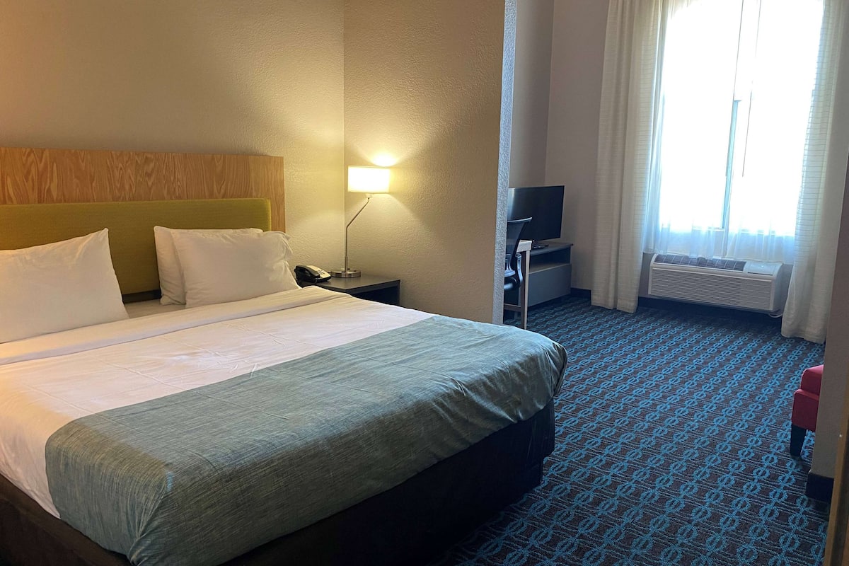 A hotel room with a bed, a desk with a lamp, a television, and an air conditioning unit.