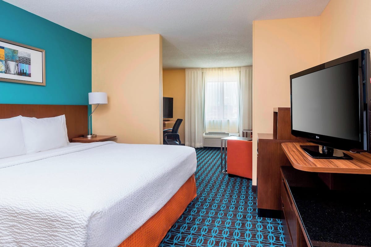 A hotel room with a large bed, a desk with a flat-screen TV, a chair, a lamp, and a window with curtains.
