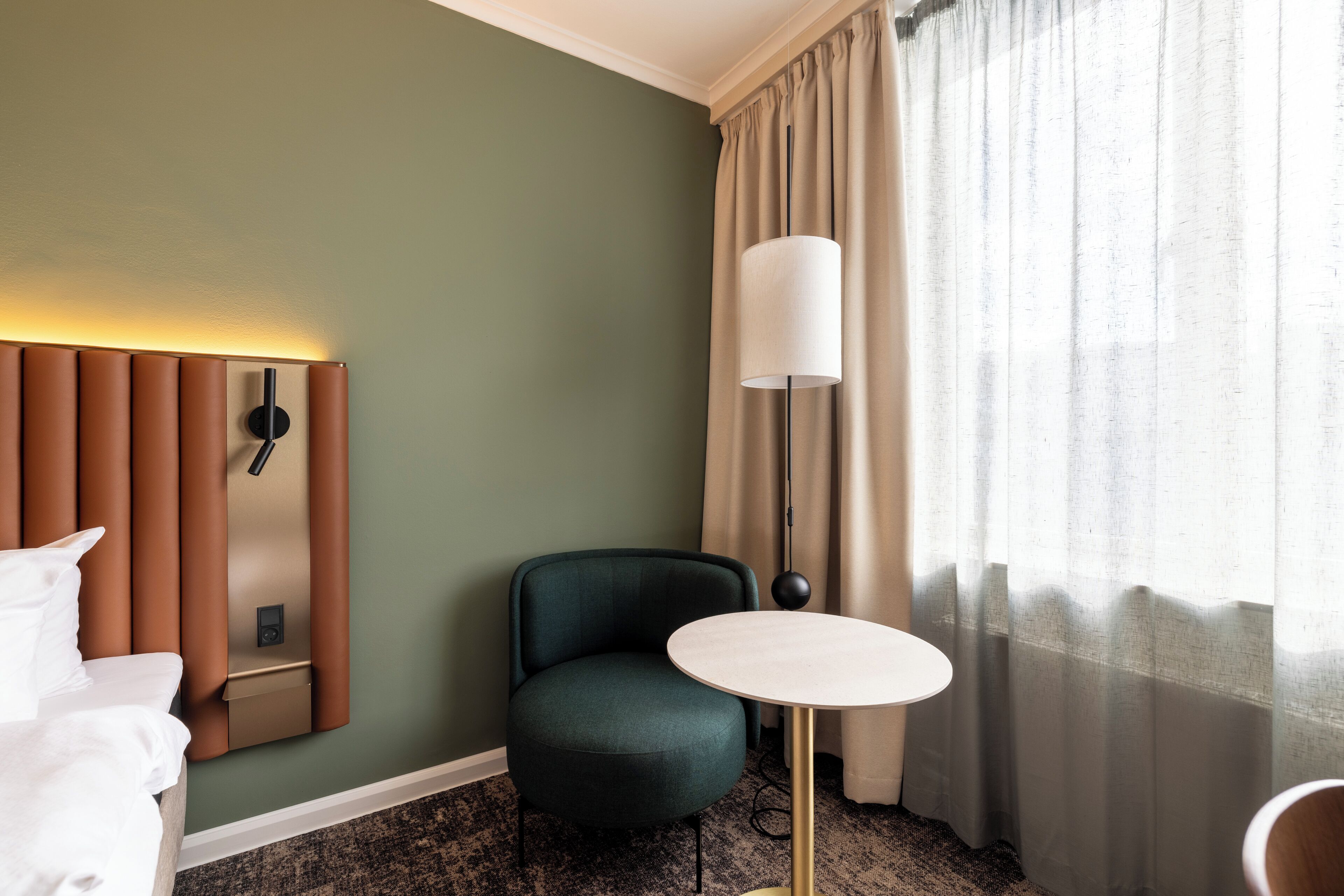 Habitación de hotel con pared verde, cabecero marrón, sillón verde, una mesa redonda, una lámpara y una ventana con cortinas.