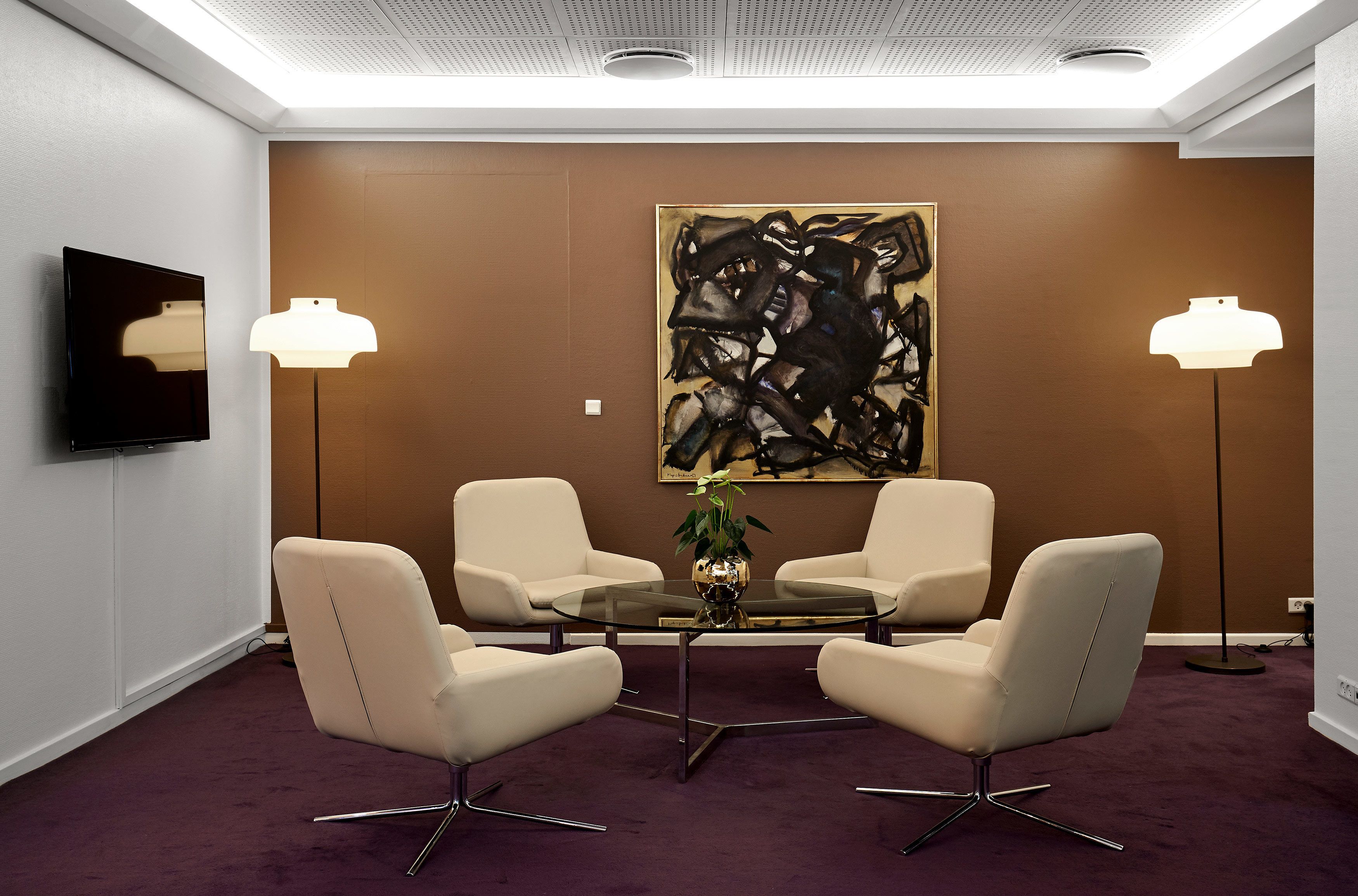 A conference room with a glass table, four chairs, a large abstract painting, two floor lamps, and a TV on the wall.