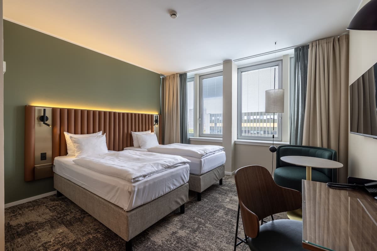 A hotel room with two beds, a desk, a chair, and large windows.