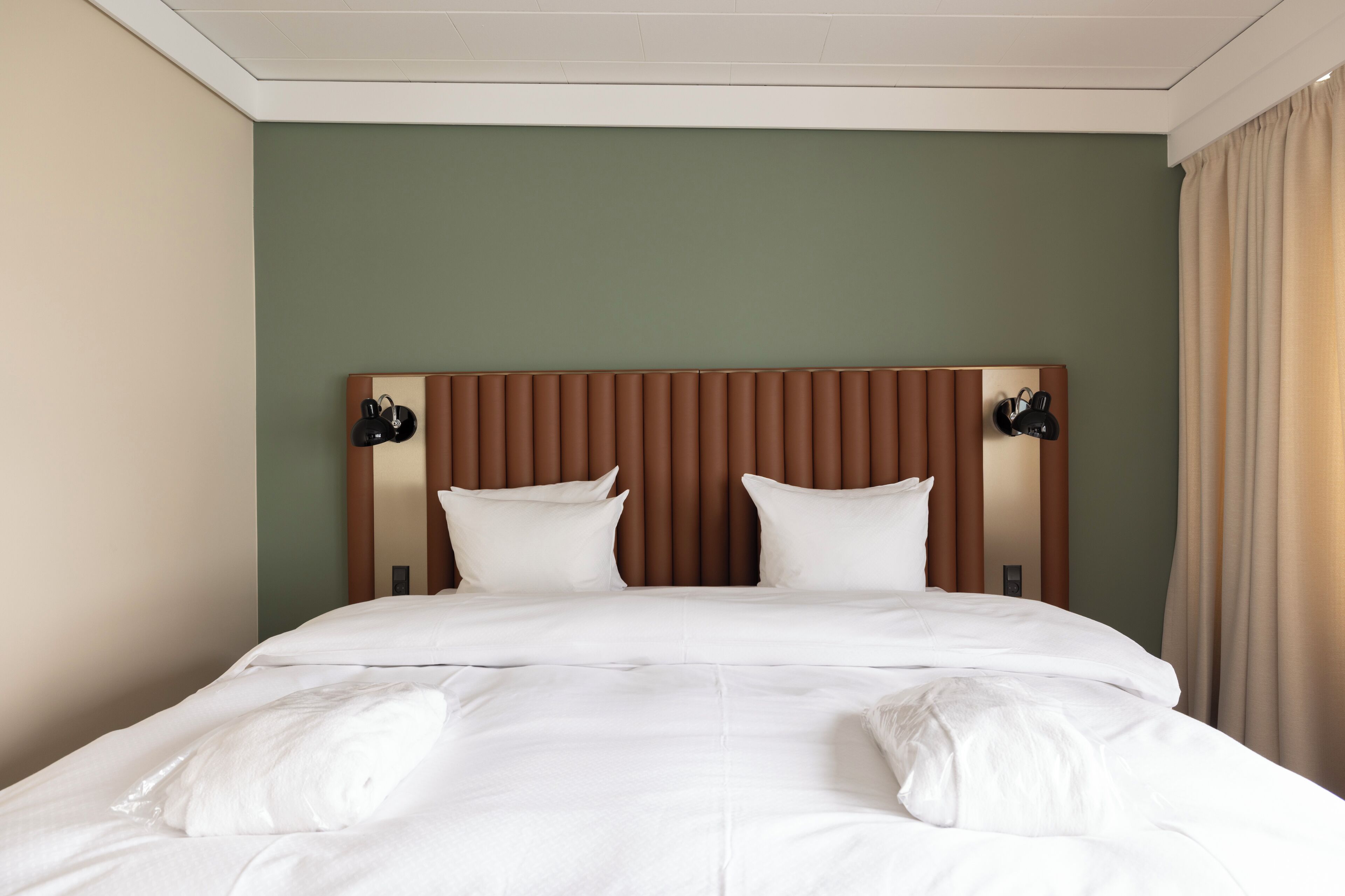 A neatly made bed with white linens and pillows, a wooden headboard, and two wall-mounted lamps.