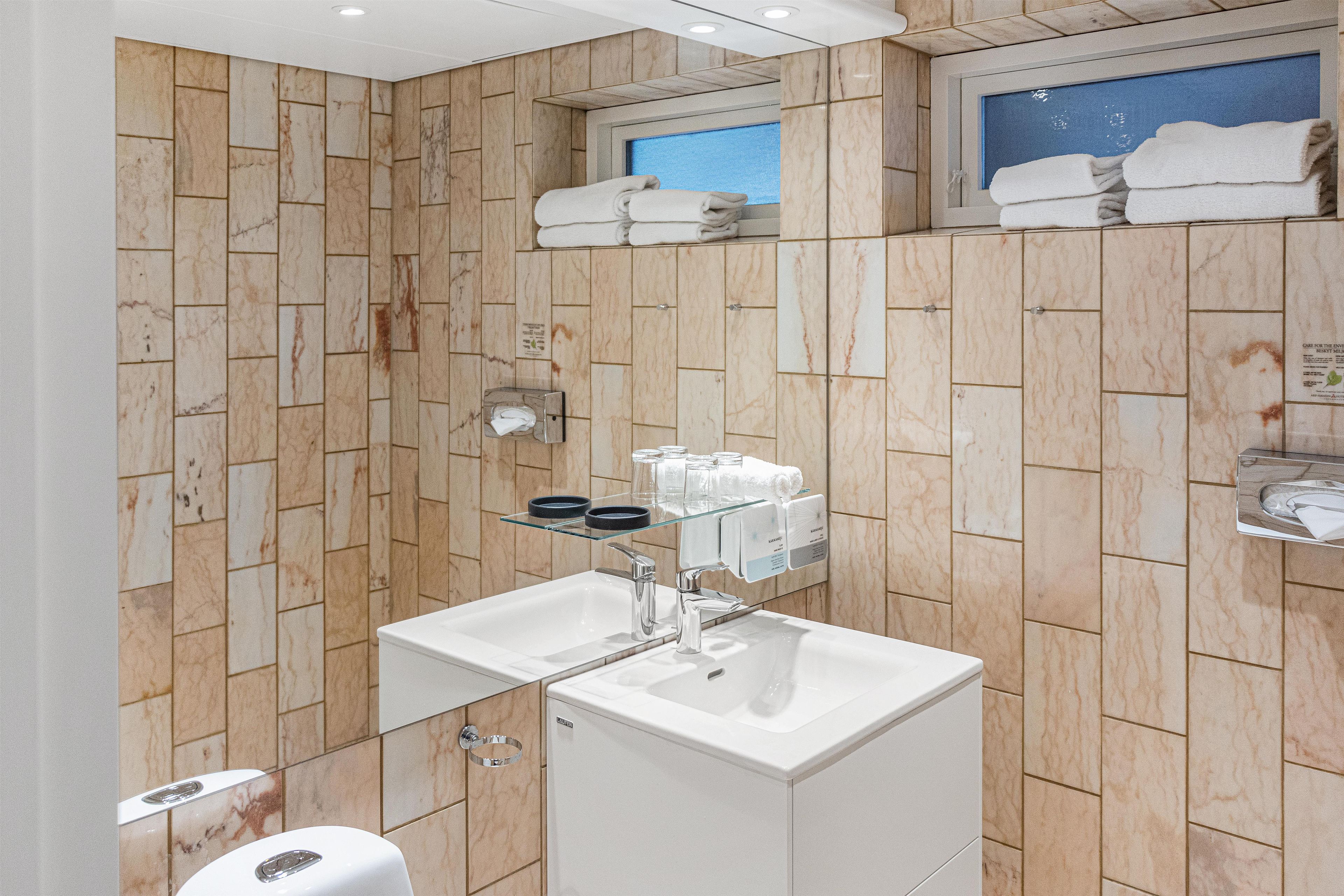 Un baño con paredes de mármol, dos lavamanos, un estante de vidrio con artículos de aseo y un perchero con toallas dobladas.