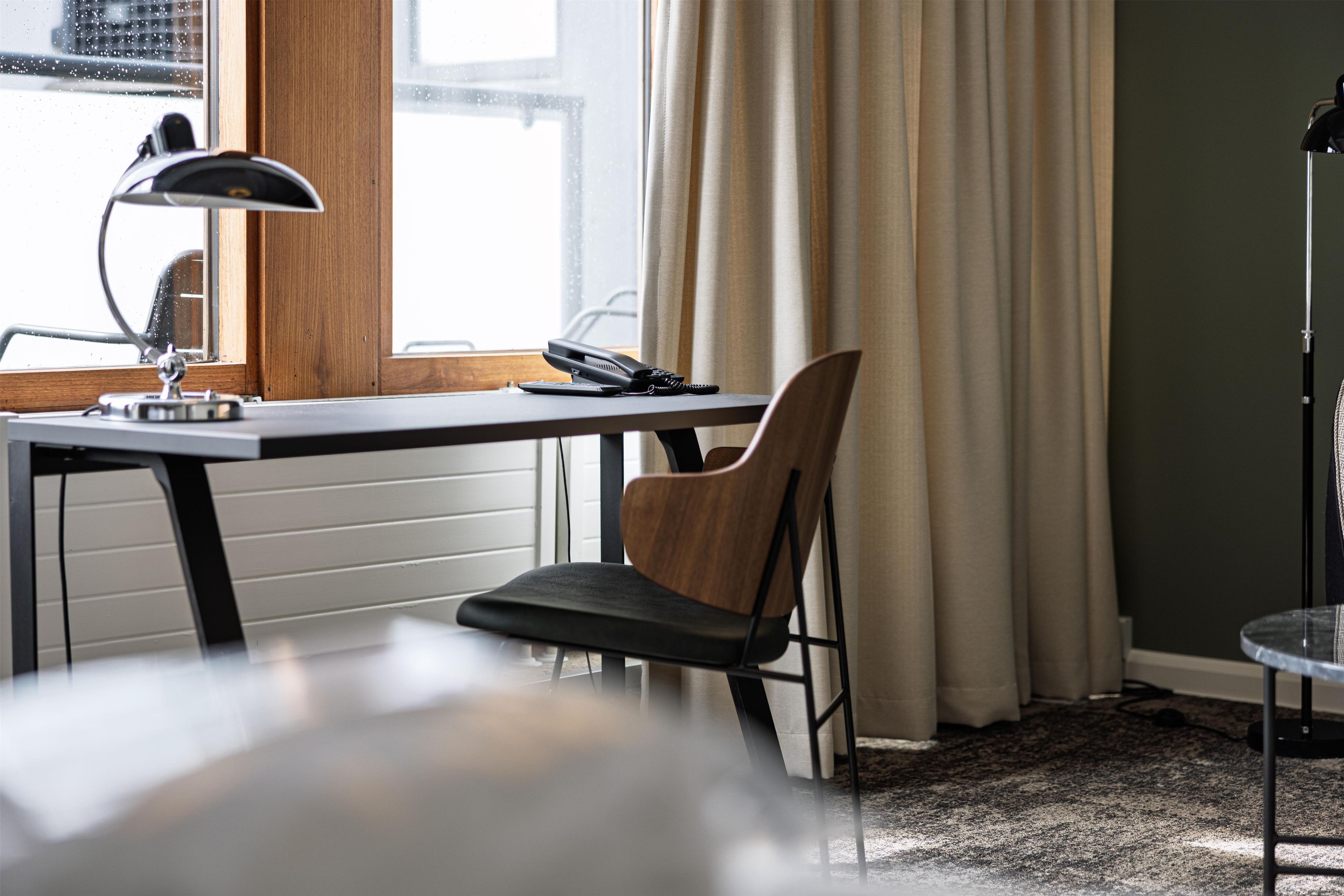Una habitación con escritorio, silla, lámpara y ventana con cortinas.