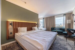 A hotel room with a large bed, a desk with a chair, a small round table, and a view of the city through large windows.