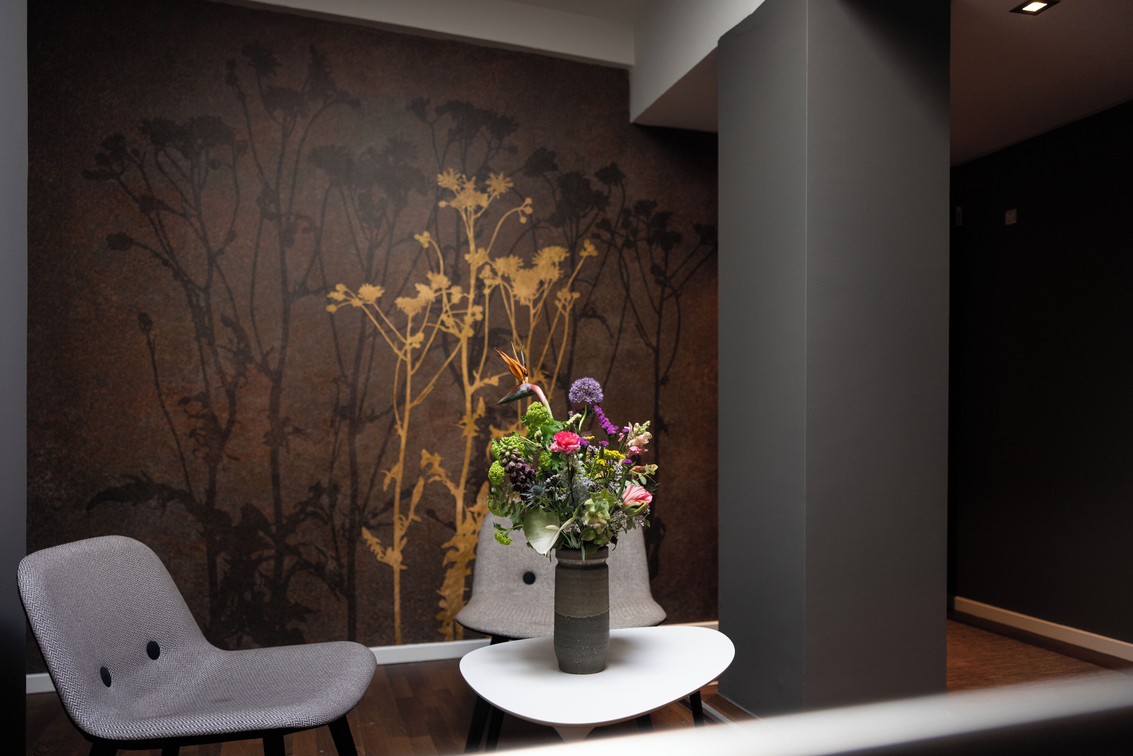 Een modern interieur met een bloemstuk aan de wand, een grijze gestoffeerde stoel en een donkere wand met een subtiel bloemenpatroon.
