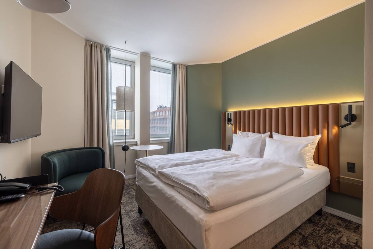 A hotel room with a bed, a desk, a chair, a TV, and a window with curtains.