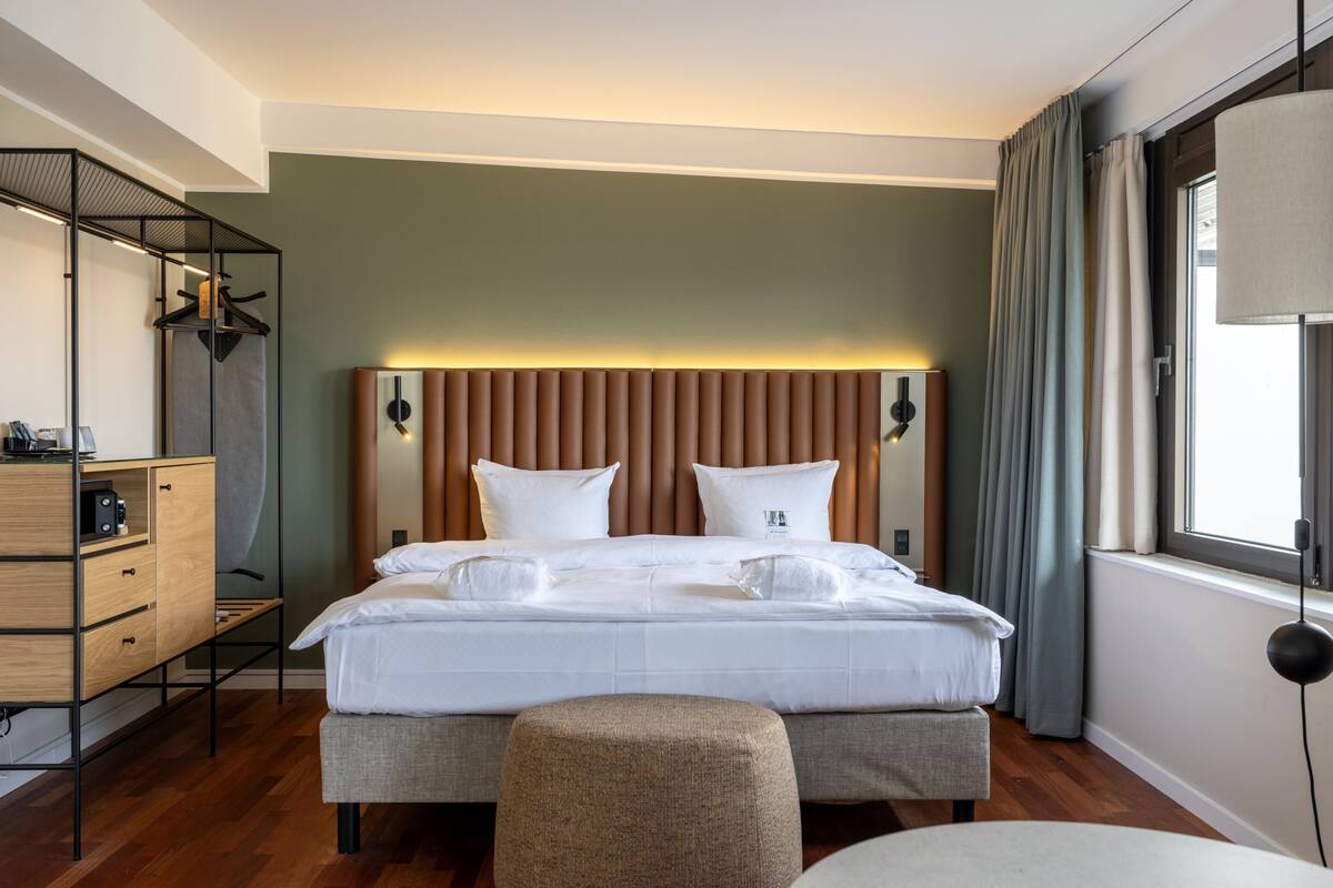 A modern hotel room with a large bed, a wooden dresser, a bedside table, and a window with curtains.