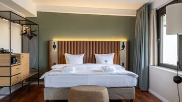 A modern hotel room with a large bed, a wooden dresser, a bedside table, and a window with curtains.