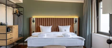 Una habitación de hotel moderna con una cama grande, un aparador de madera, una mesita de noche y una ventana con cortinas.