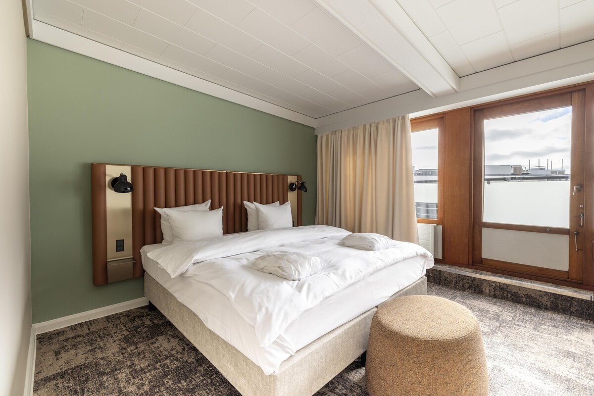 Habitación de hotel con una cama grande, un reposapiés redondo, vista al agua a través de la ventana y un cabecero de madera.