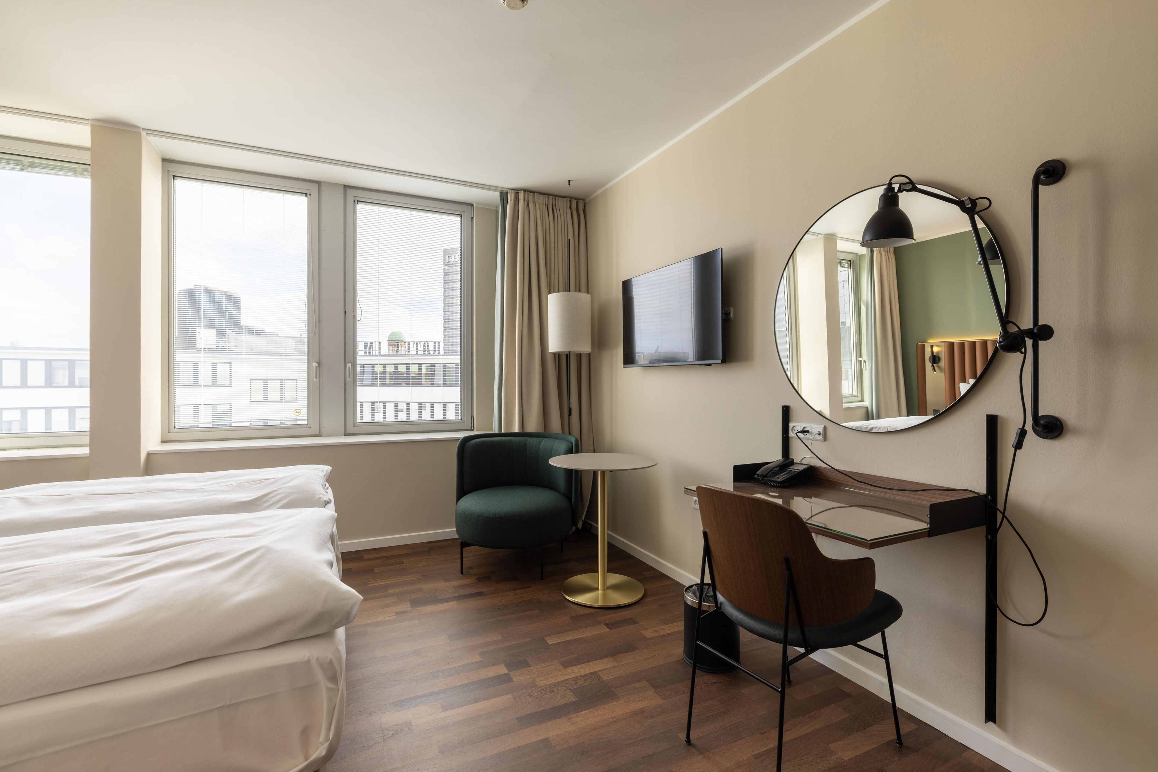 Quarto de hotel com cama, escrivaninha, cadeira, espelho redondo e televisão.