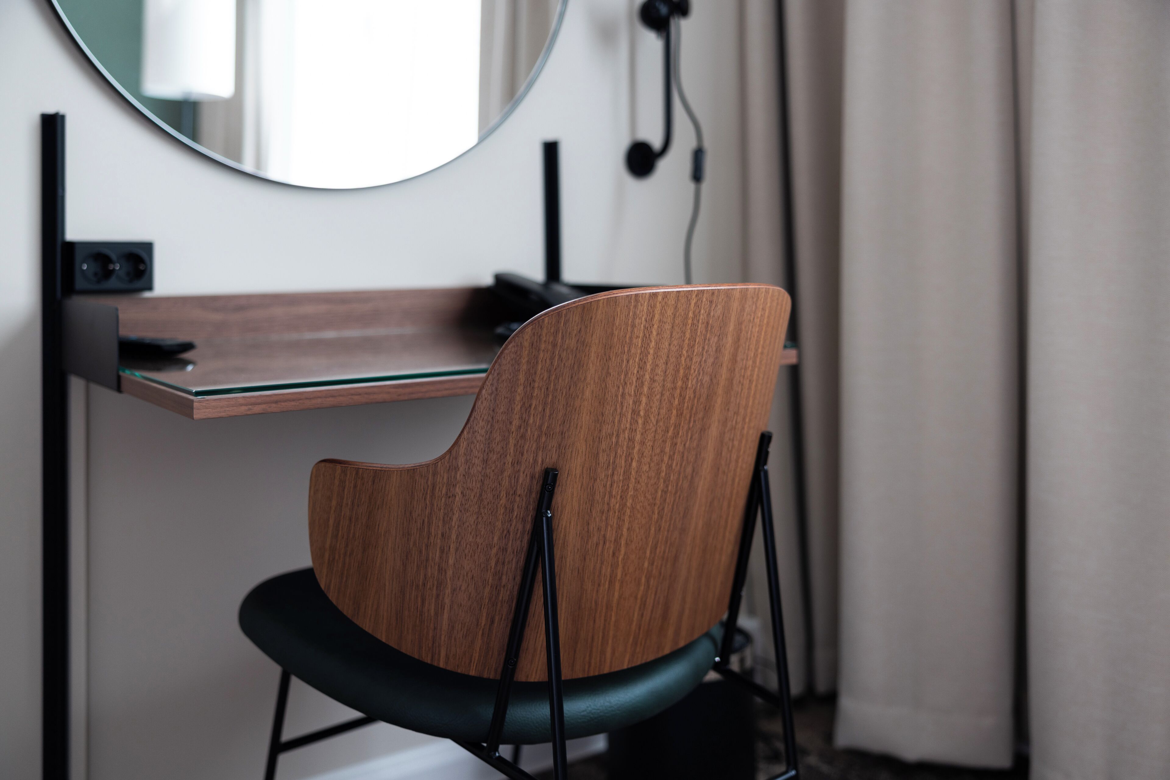 A modern chair with a wooden backrest and black seat, positioned in front of a desk with a glass top and a round mirror above it.