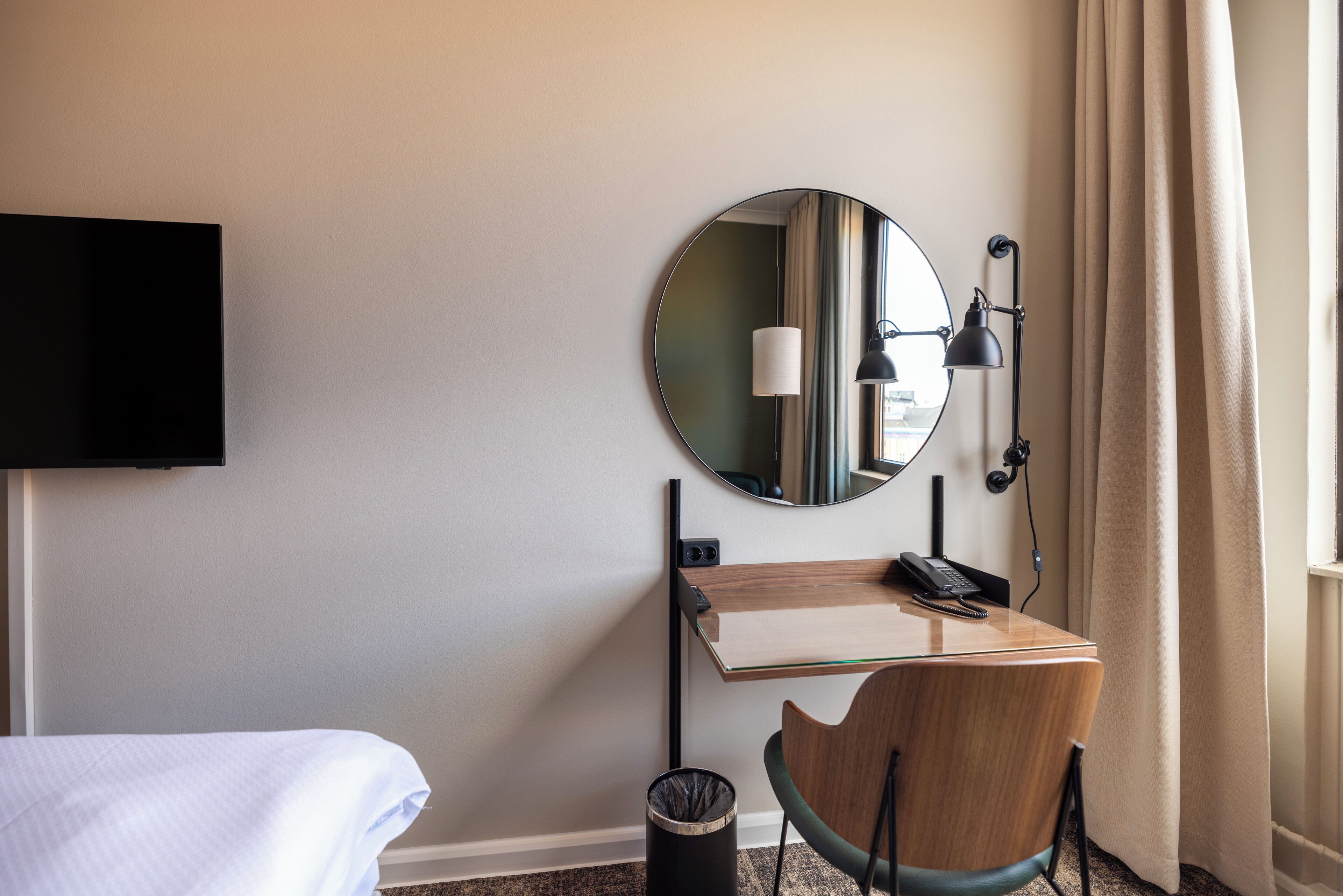 A hotel room with a bed, a desk with a telephone, a mirror, a lamp, and a trash can.