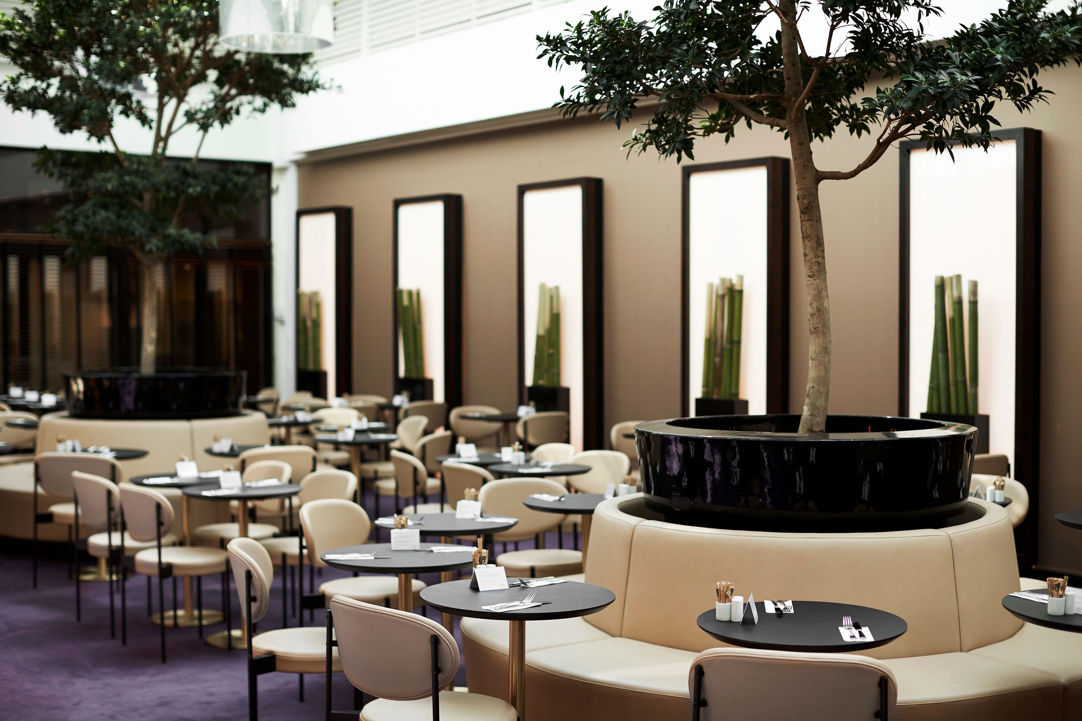 Un moderno vestíbulo de hotel con mesas redondas, sofás color beige y una jardinera negra central con un árbol.