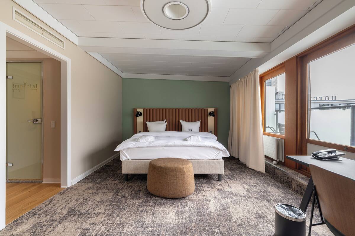 A hotel room with a bed, a small ottoman, a desk, and a view of the city through the window.