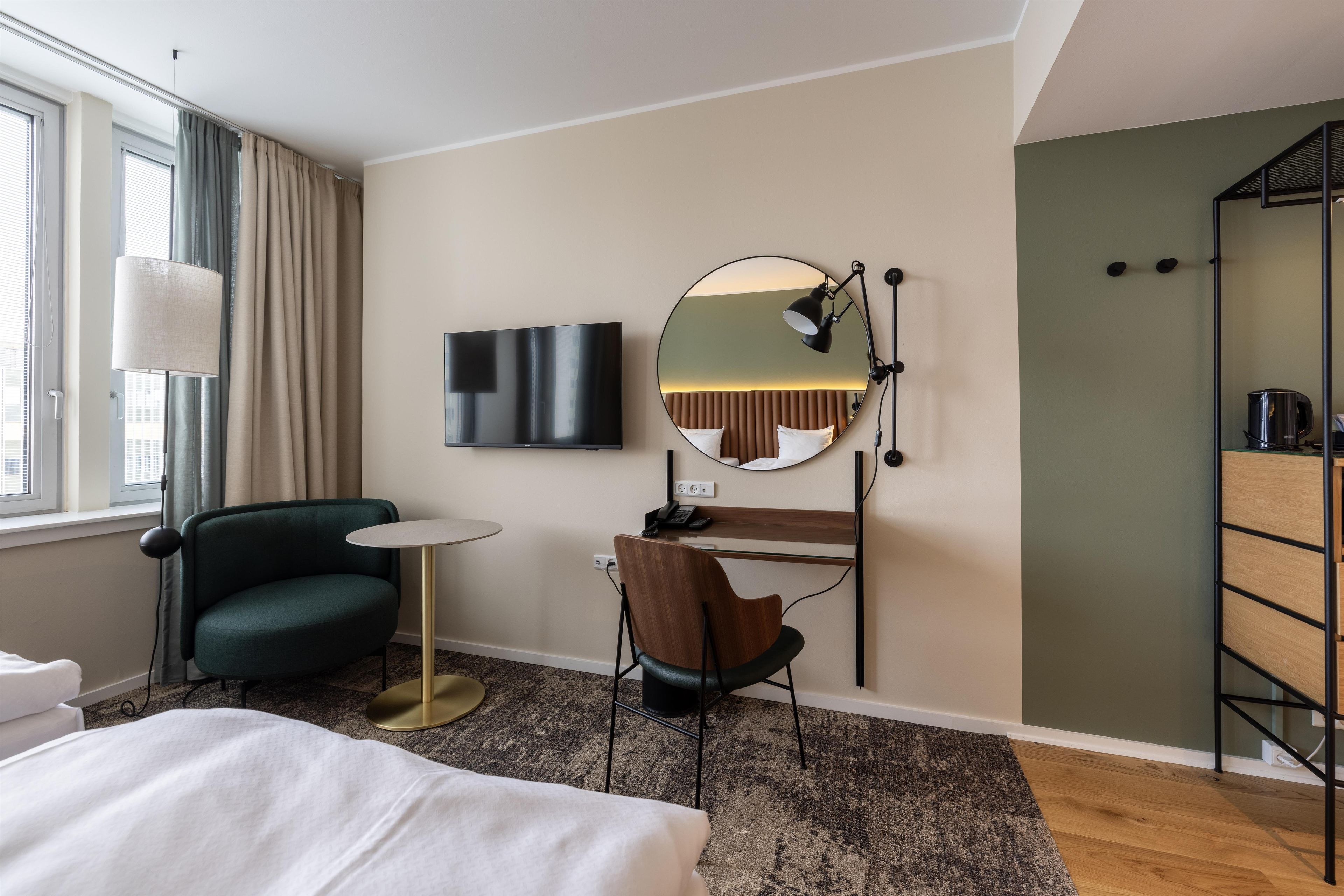 A hotel room with a bed, a desk, a chair, a round mirror, a television, and a window with curtains.