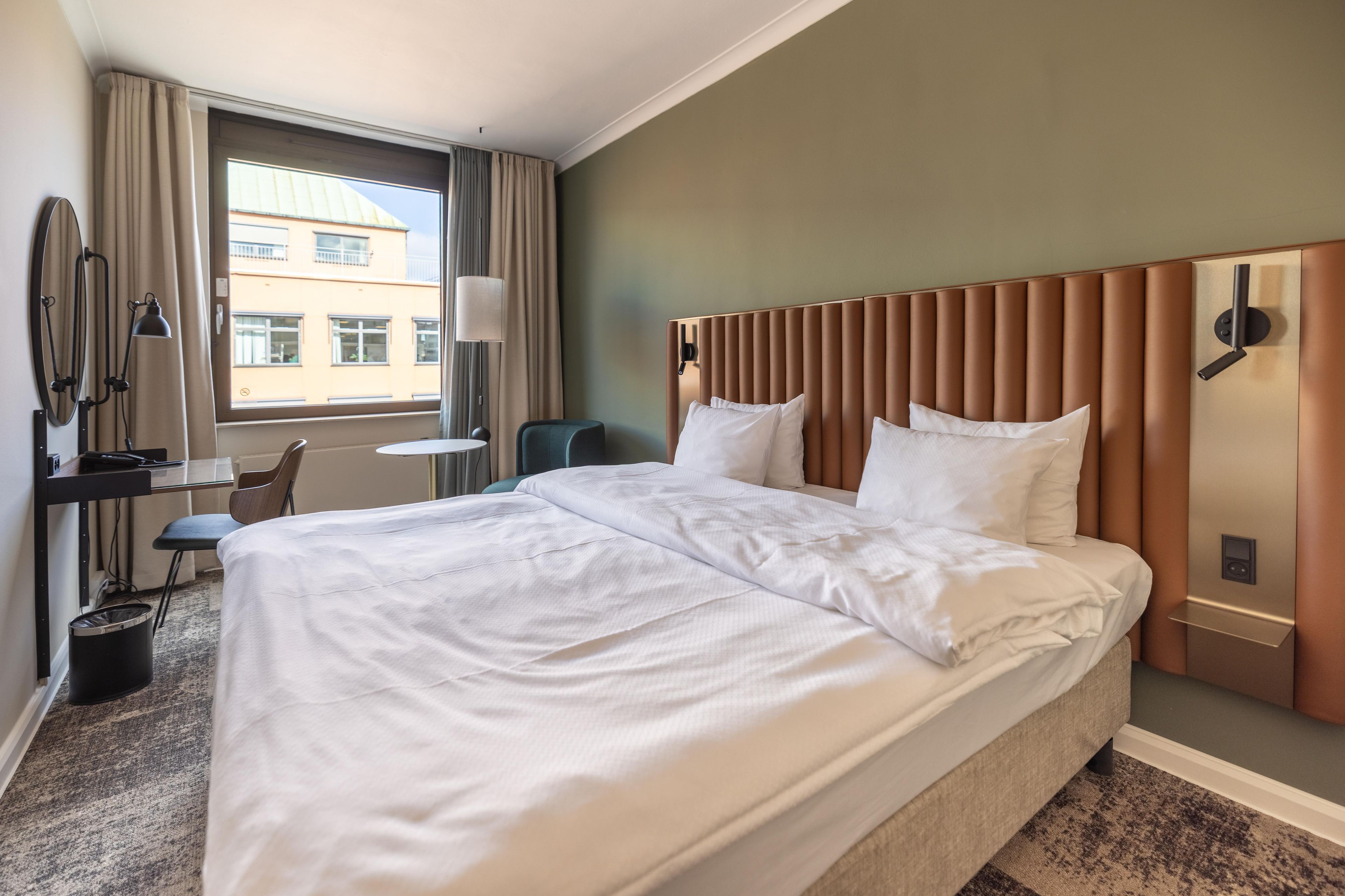A hotel room with a large bed, a desk with a chair, a lamp, and a view of buildings outside the window.