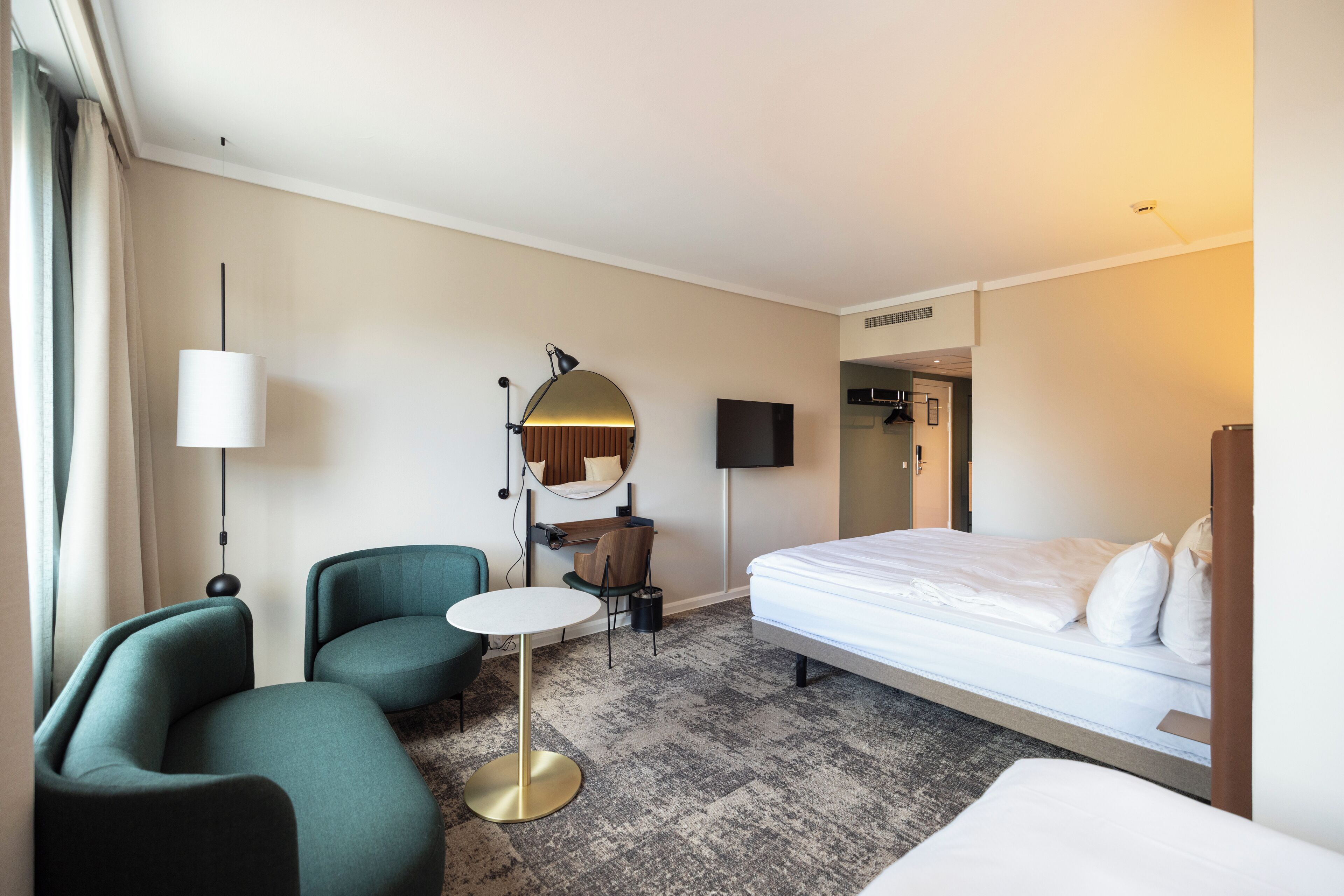 A hotel room with a bed, two armchairs, a small round table, a lamp, a TV, and a mirror.