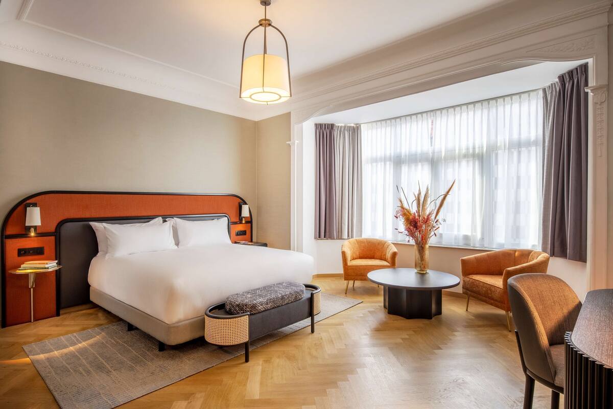 A hotel room with a large bed, a bedside table, a lamp, a chair, a round table, and a vase with dried plants.