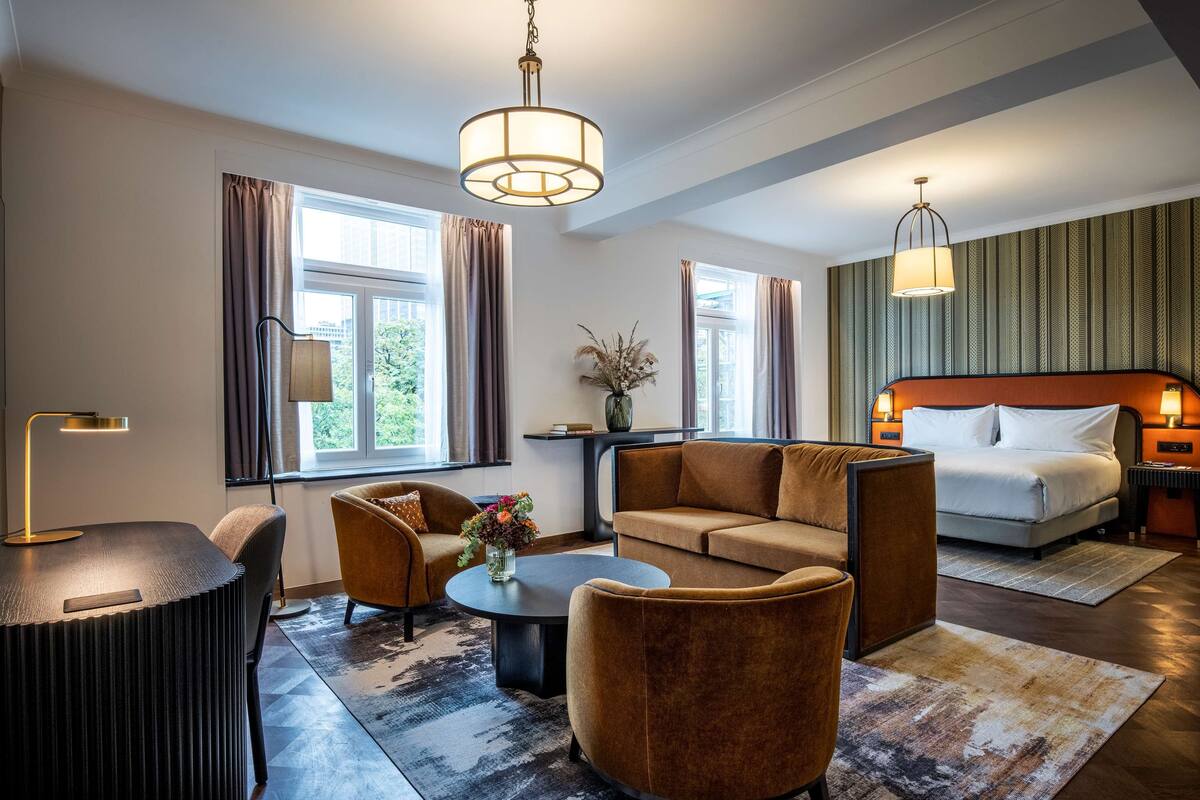 A modern hotel room with a sofa, armchair, coffee table, and a bed with a headboard.