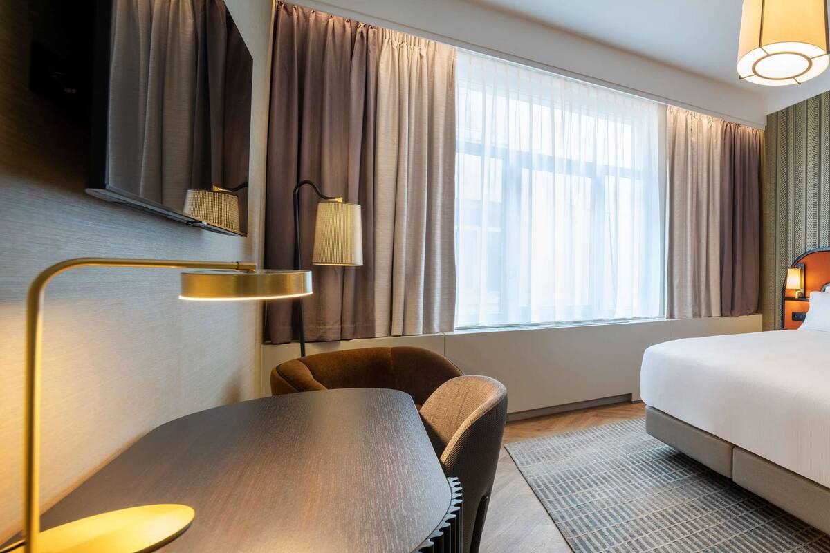 A hotel room with a bed, a desk, a chair, a lamp, and a window with curtains.