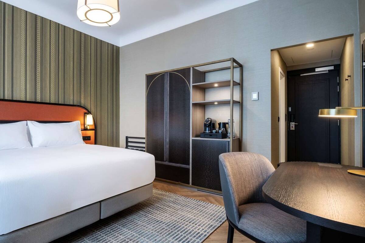 A hotel room with a bed, bedside tables, a desk, a chair, a wardrobe, and a mirror.