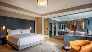A modern hotel room with a large bed, a sitting area with a chair, a small table, and a glass-enclosed bathroom with a bathtub. - DoubleTree by Hilton Brussels City (Brussels)