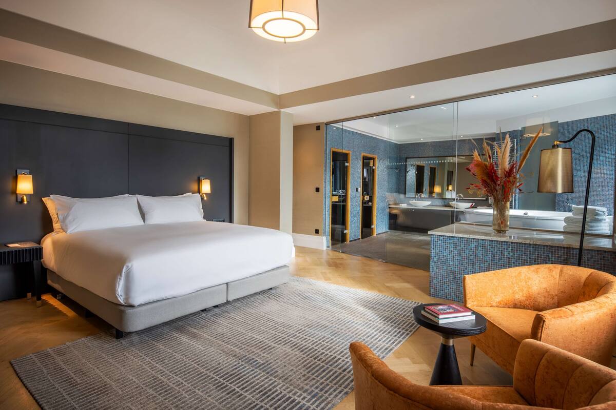A modern hotel room with a large bed, a sitting area with a chair, a small table, and a glass-enclosed bathroom with a bathtub.