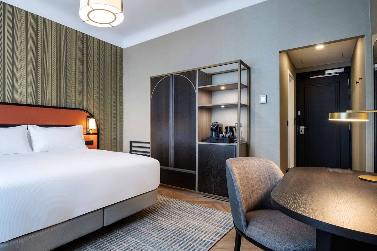 A hotel room with a bed, bedside tables, a desk, a chair, a wardrobe, and a mirror.