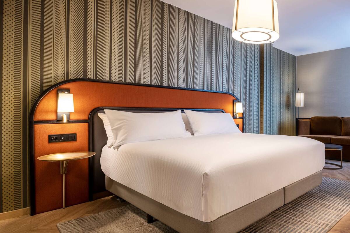 A hotel room with a large bed, a desk, and a chair. There are wall sconces on either side of the bed and a lamp on the desk.