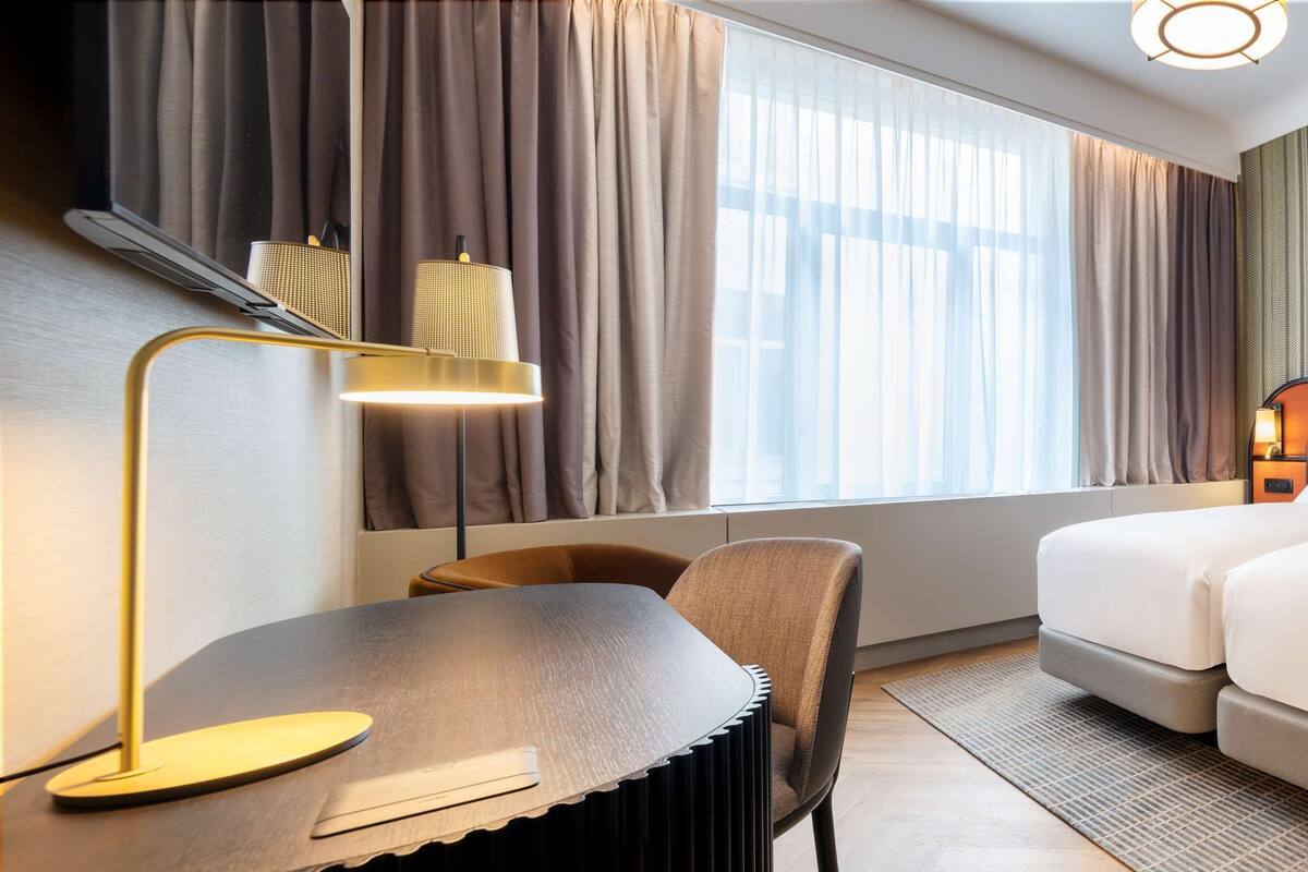 A hotel room with a bed, a desk, a chair, and a window with curtains.