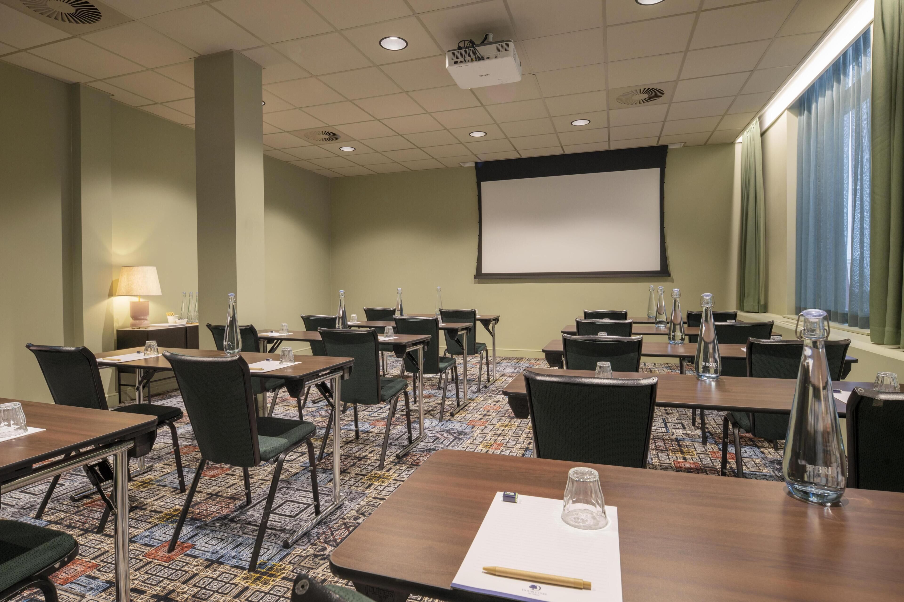 Une salle de conférence équipée de tables, de chaises, d’un écran de projection et d’une grande fenêtre avec des rideaux.