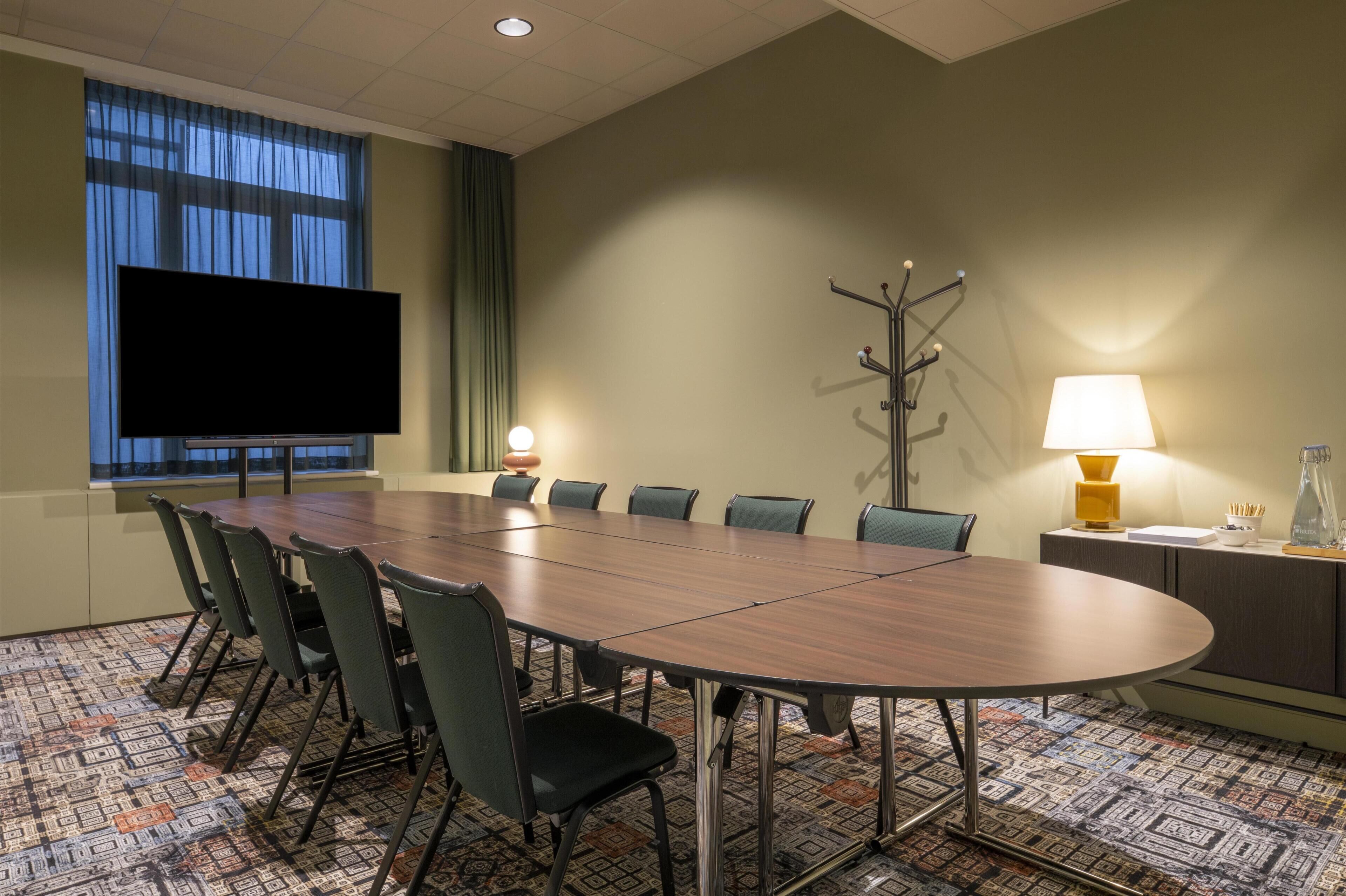A conference room with a long table, chairs, a TV screen, a coat rack, a lamp, and a patterned carpet.