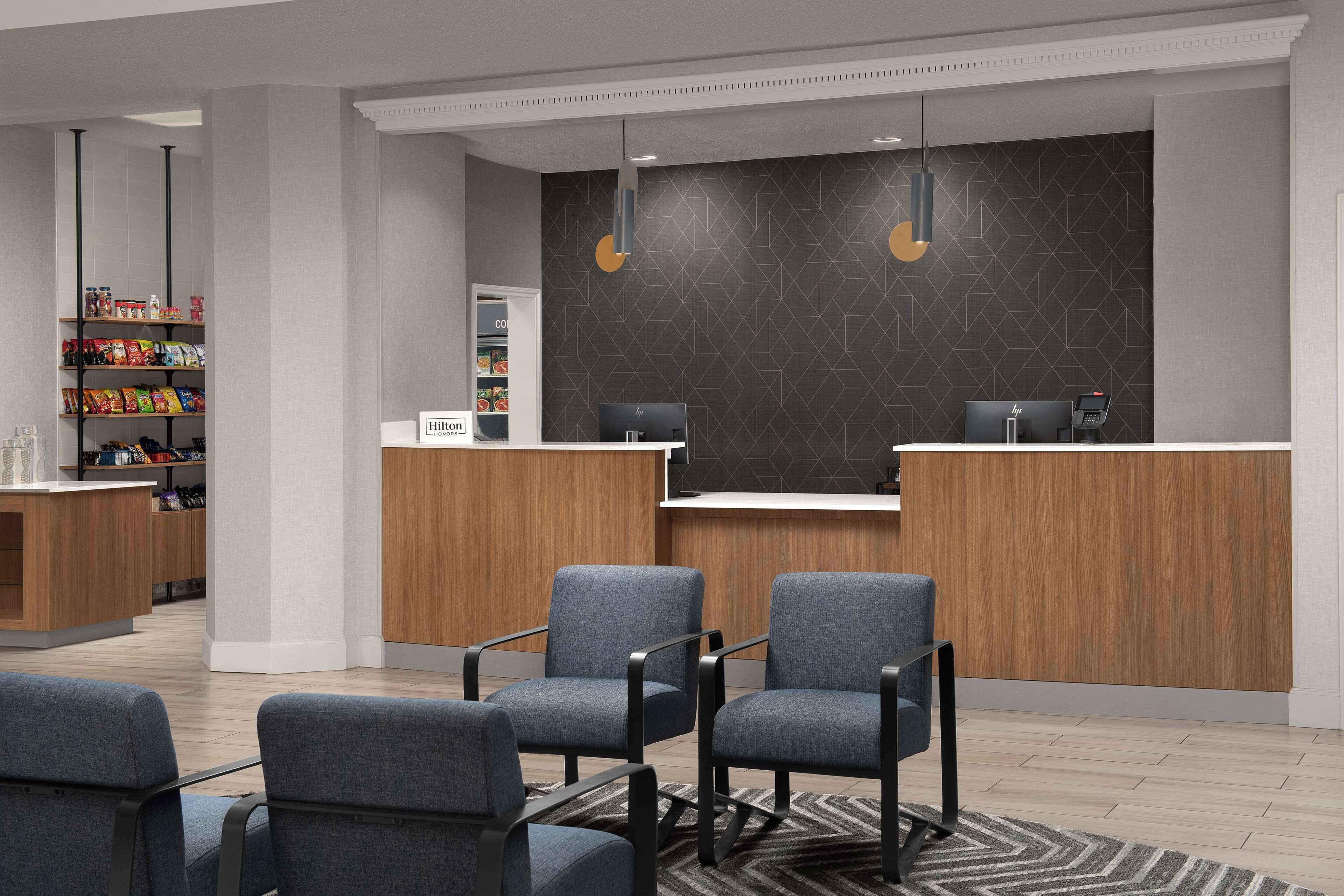 Un área de recepción moderna con un mostrador de madera, dos sillas grises, un estante con aperitivos y una pared de color oscuro con motivos.