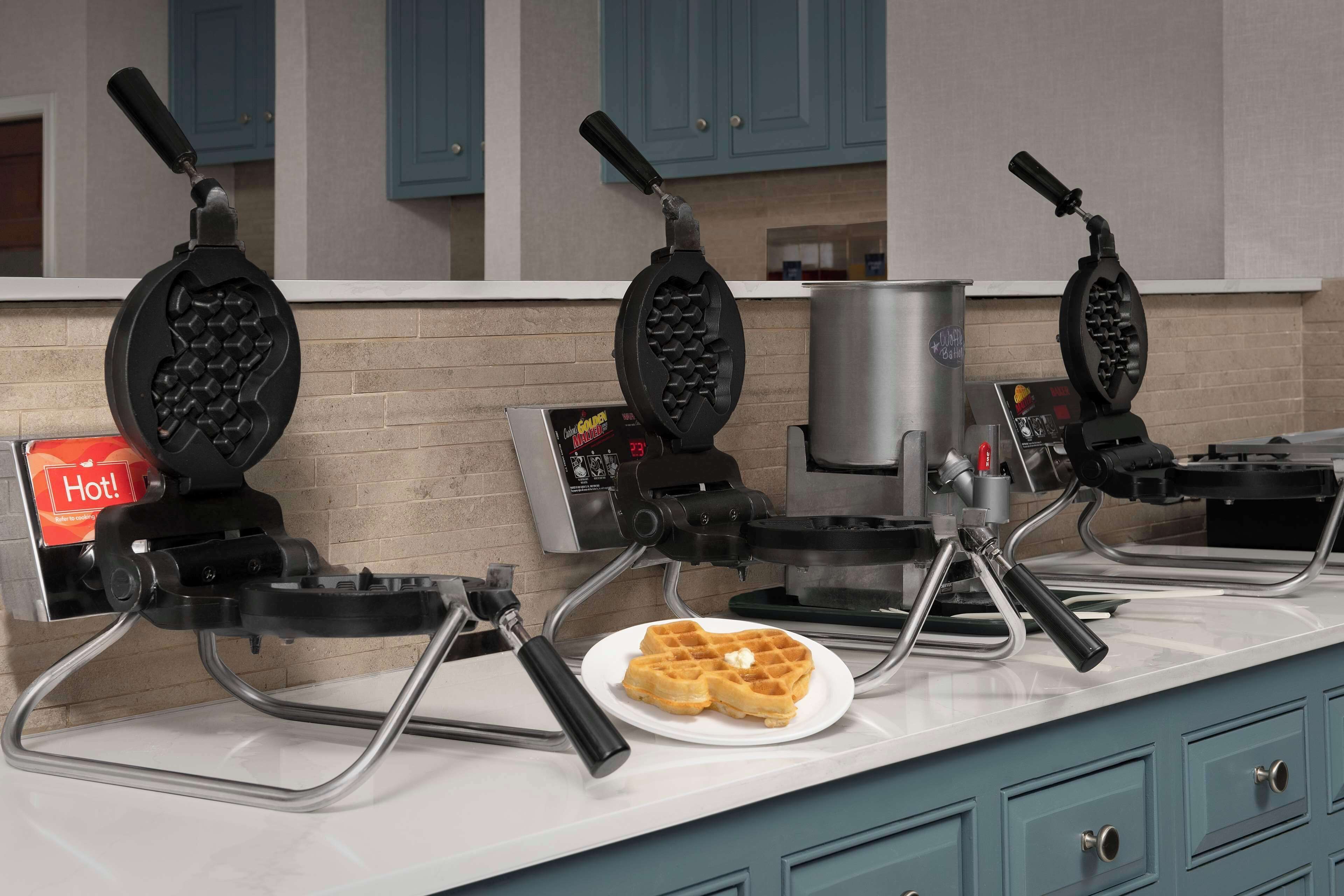 Três máquinas de waffle sobre uma bancada de cozinha, com um prato de waffles e um aviso que diz 'Quente!'.
