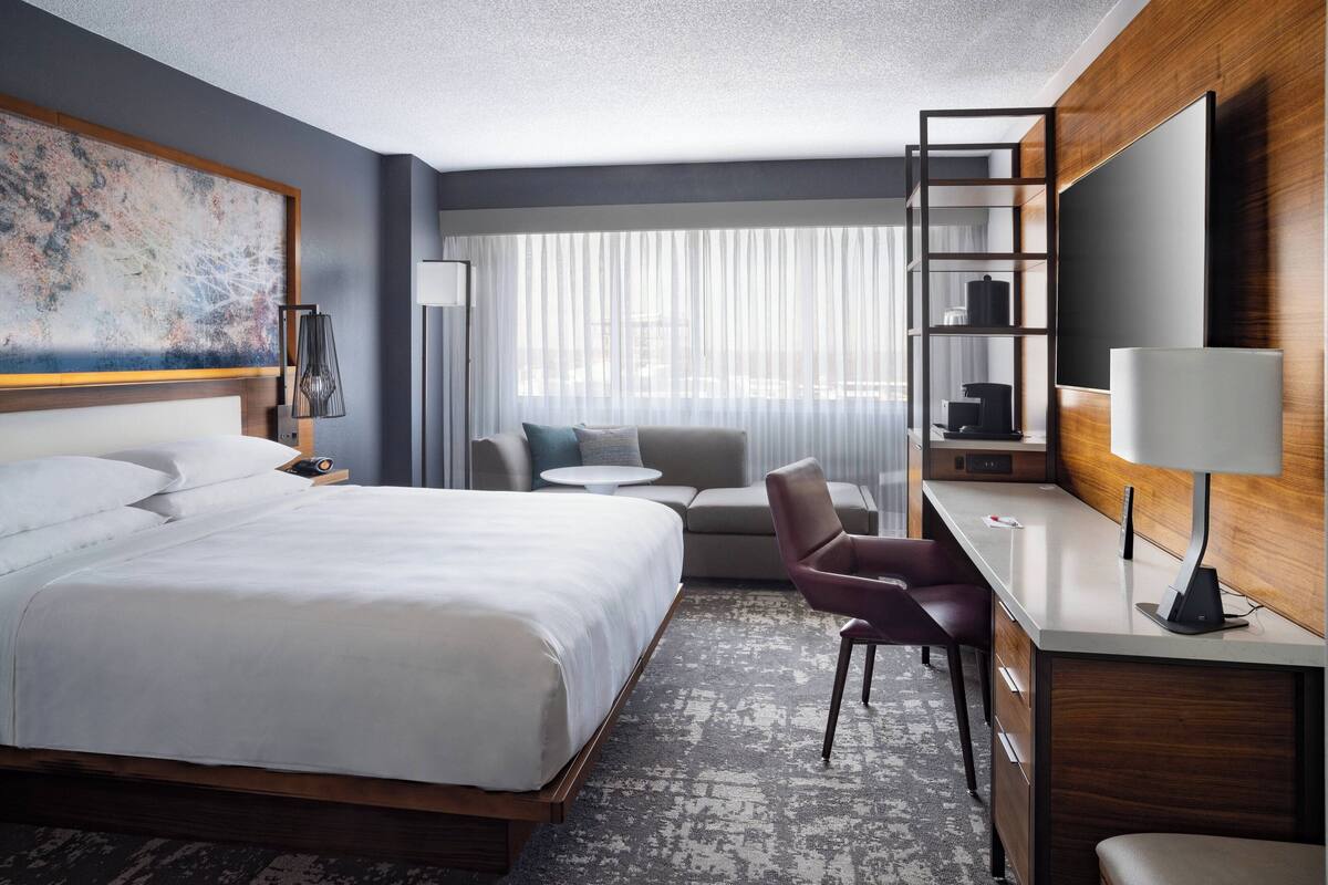 A modern hotel room with a large bed, a desk with a chair, a sofa, a TV, and a large window with sheer curtains.