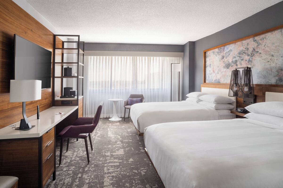 A hotel room with a desk, two beds, a TV, a chair, a small table, and a large window with sheer curtains.