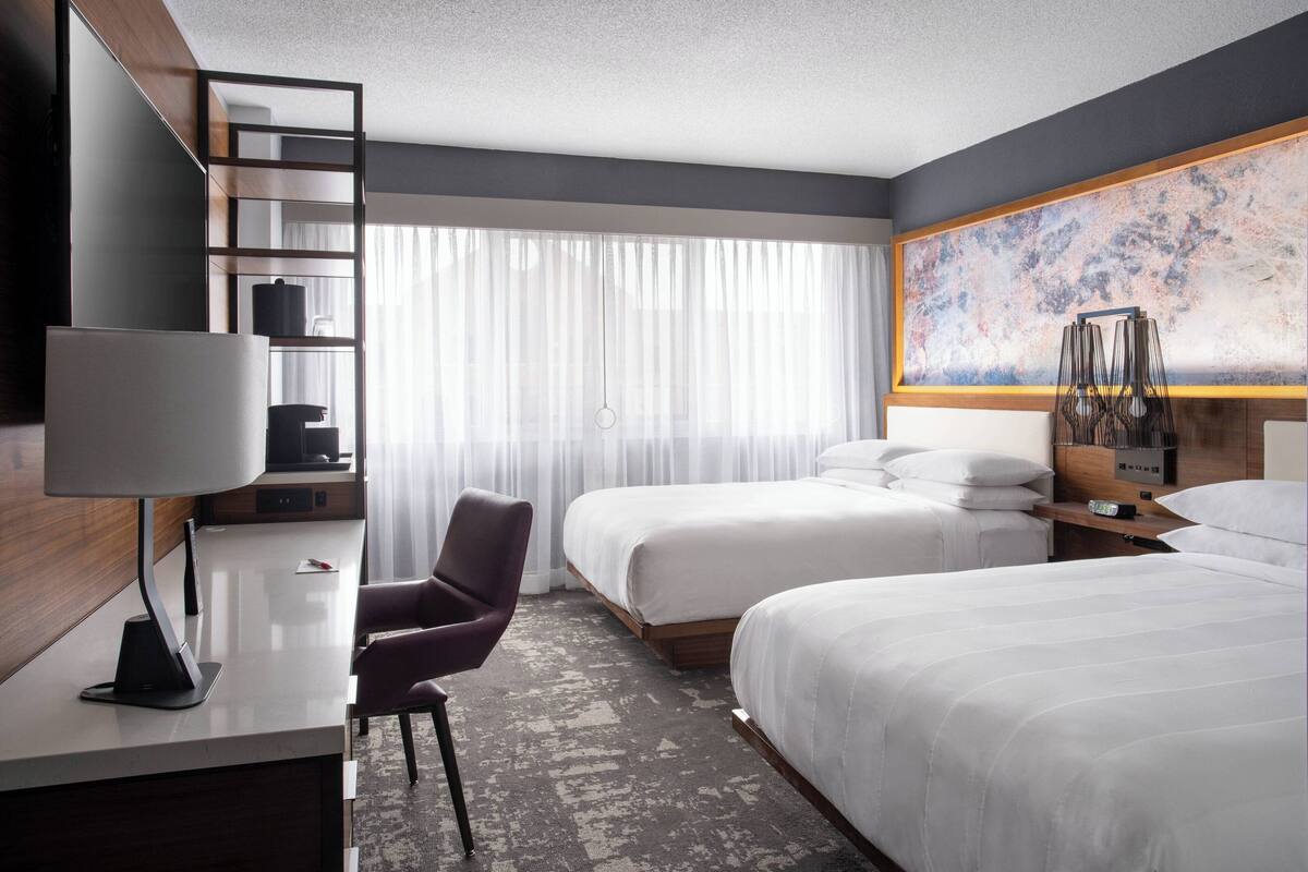 A hotel room with two beds, a desk with a lamp, a chair, a bookshelf, and a large abstract painting on the wall.