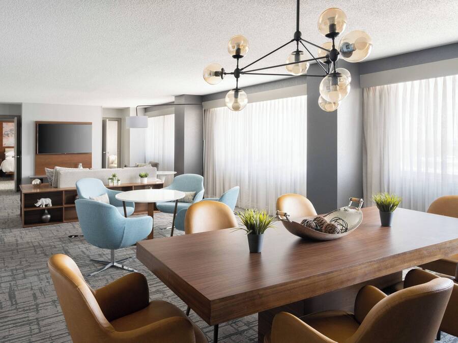 A modern hotel room with a dining area, a sofa set, and a TV mounted on the wall.