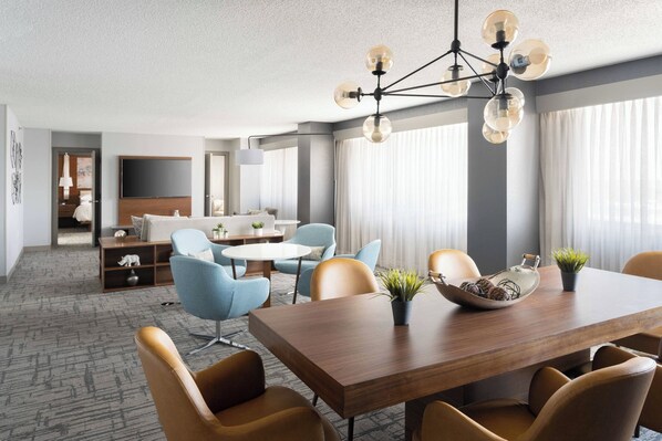 A modern hotel room with a dining area, a sofa set, and a TV mounted on the wall.