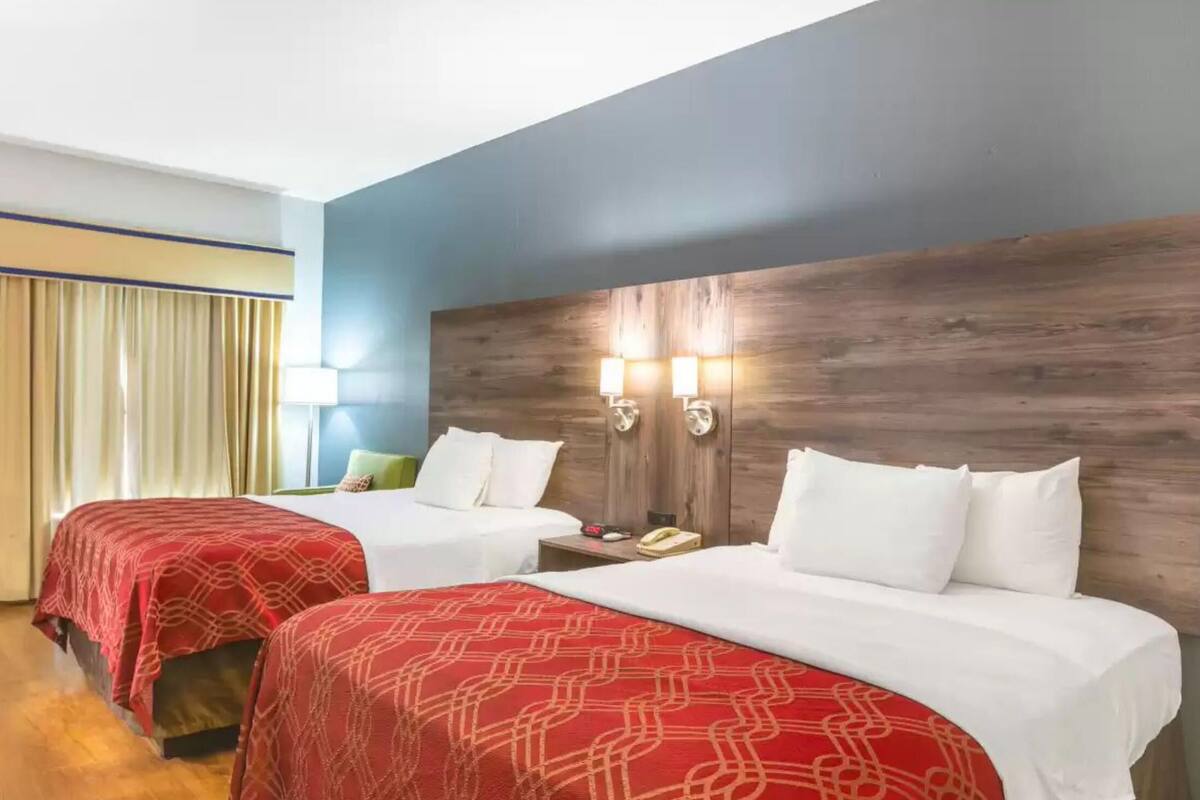 A hotel room with two beds, wooden headboard, bedside lamps, and a window with curtains.