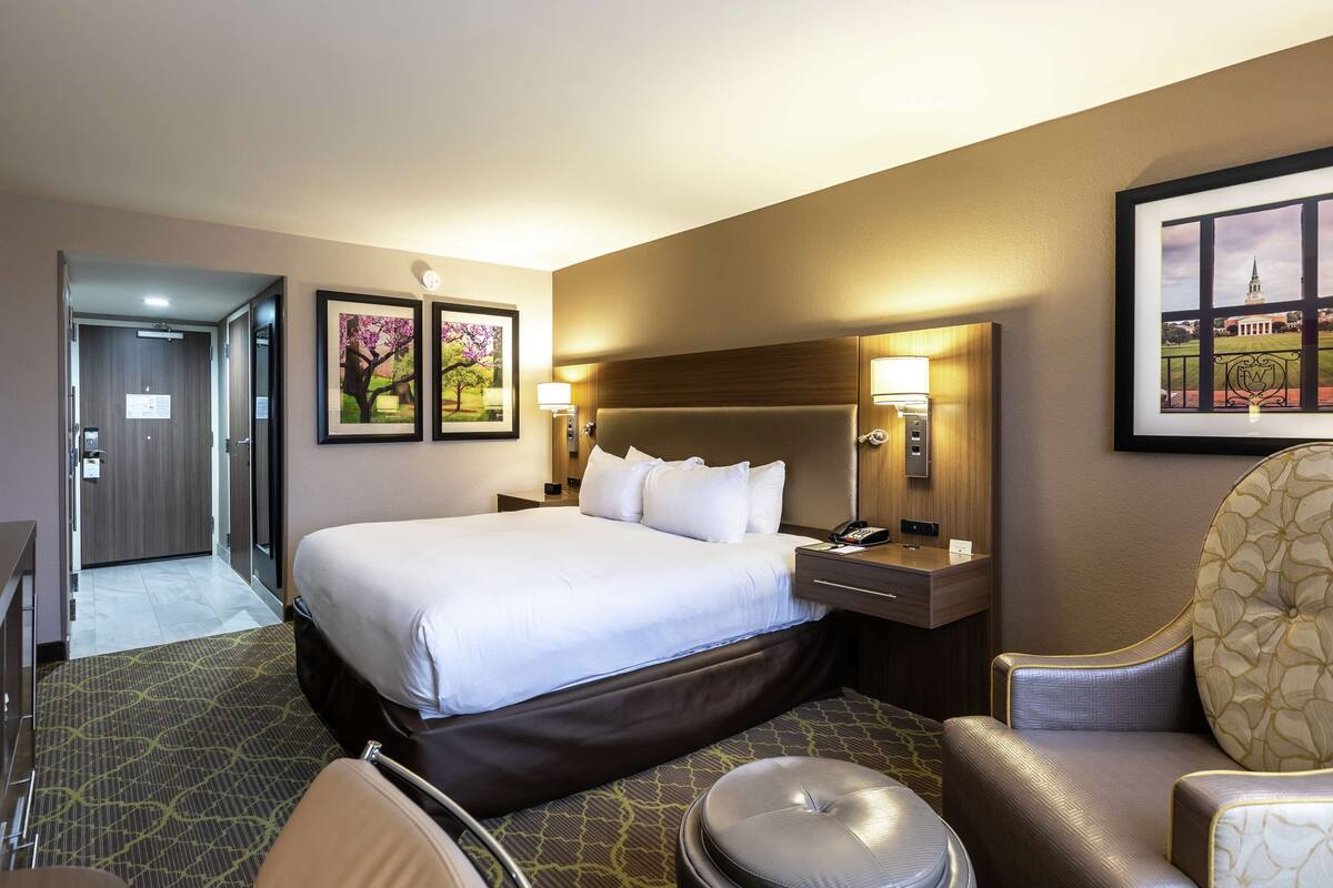 A hotel room with a large bed, a desk, a chair, and a small table. There are framed pictures on the wall and a lamp on the desk.