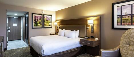 A hotel room with a large bed, a desk, a chair, and a small table. There are framed pictures on the wall and a lamp on the desk.