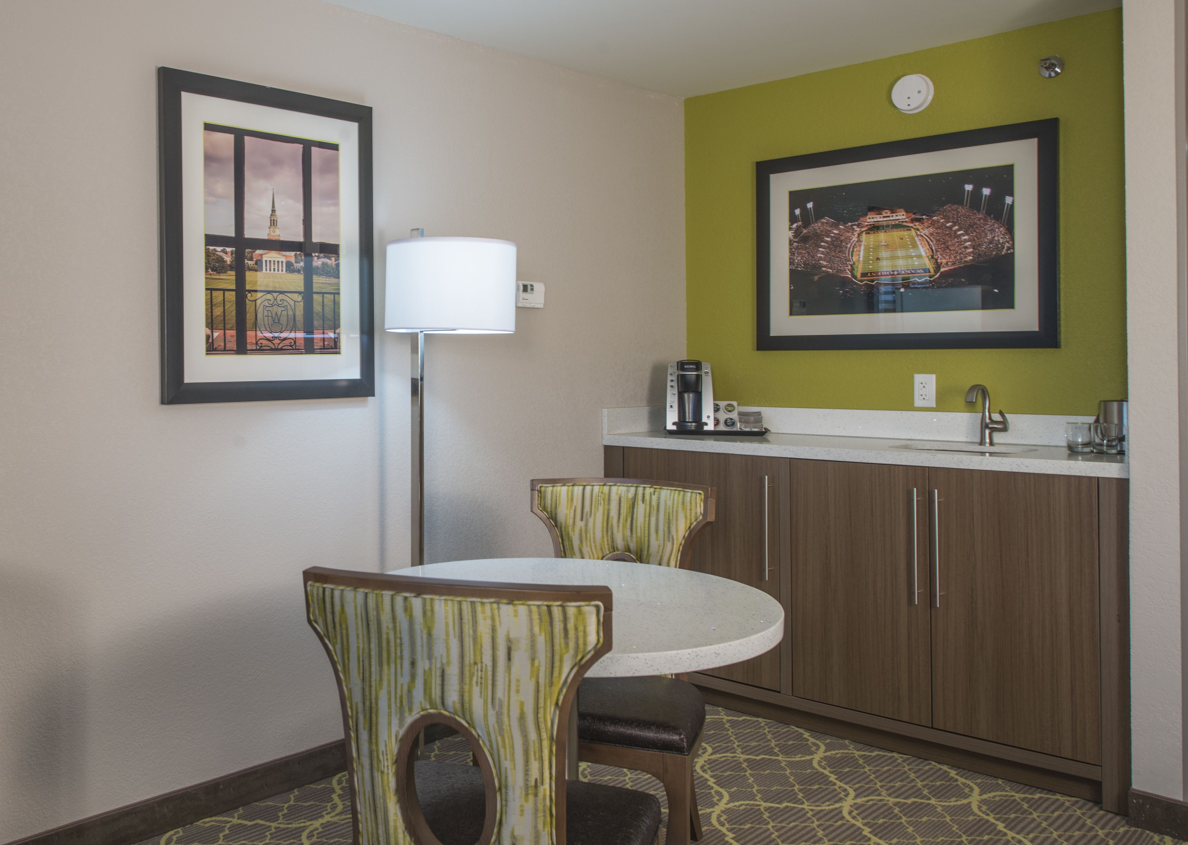 A hotel room with a round dining table, two chairs, a floor lamp, a coffee maker, and a framed picture of a stadium on the wall.